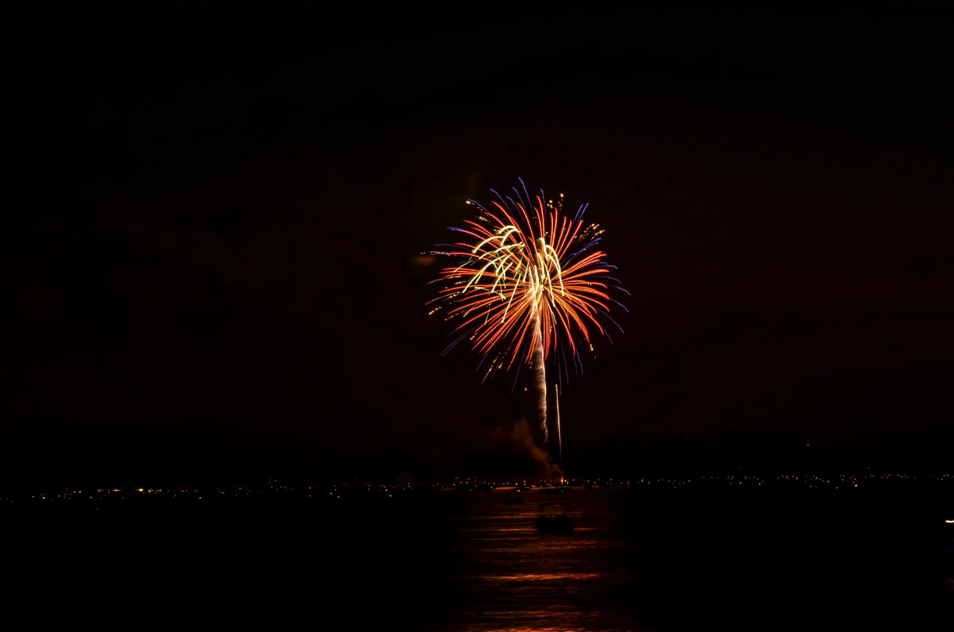 Fireworks bursting over open water at night, with boats gathered below and shoreline lights along the far bank.